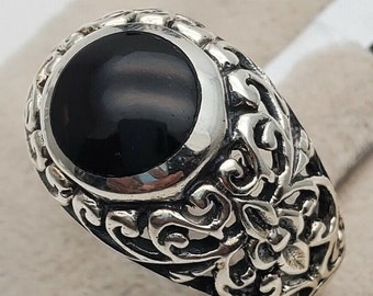 May include: A silver ring featuring a large, smooth, circular black stone set in the center. The band is crafted with intricate, raised scrollwork and filigree patterns that extend around the sides of the ring, creating a textured, metallic finish.
