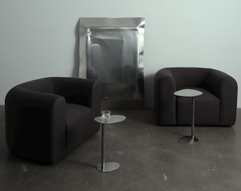 May include: Two black armchairs with rounded edges and a silver metal side table with a glass on top. A large silver metal sheet is in the background.