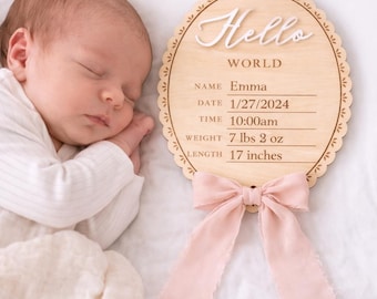 May include: An oval wooden birth announcement sign with a scalloped edge and a light pink fabric bow. The sign features the white 3D text Hello, the word WORLD, and printed lines for Name Emma, Date 1/27/2024, Time 10:00am, Weight 7 lbs 2 oz, and Length 17 inches.