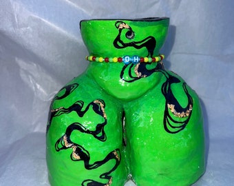 May include: A green ceramic vase with black and gold swirl designs. The vase has a beaded necklace with the letters "D-O-H" around the middle.