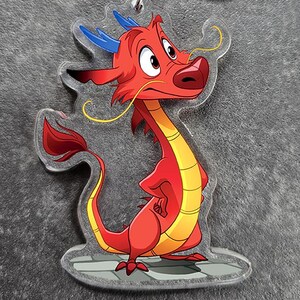 Puede incluir: Un llavero de dragón rojo y amarillo con un llavero plateado. El dragón tiene cuernos azules y detalles amarillos. El llavero es de acrílico transparente y es un personaje de dibujos animados.