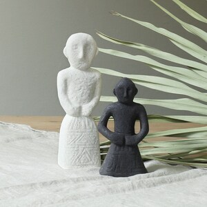 Könnte beinhalten: Zwei kleine Keramikfiguren, eine weiße und eine schwarze, stehen auf einer weißen Leinen-Tischdecke. Hinter den Figuren befindet sich ein getrocknetes Palmblatt.