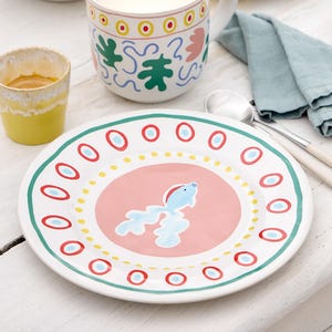 May include: A white ceramic plate with a pink centre and a blue fish design. The plate has a green, red, and yellow border with a repeating circle pattern.