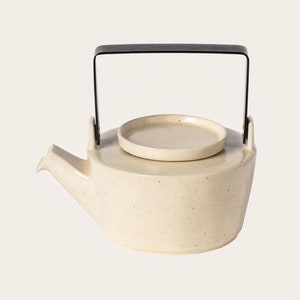 Op de afbeelding: Crèmekleurige keramische theepot met een zwart metalen handvat en een bijpassend deksel. De theepot heeft een tuit om uit te schenken en een gespikkelde textuur. Het ontwerp is minimalistisch en modern.