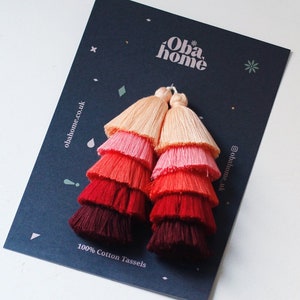 Puede incluir: Un par de pendientes de borla multicolores con un degradado de melocotón, rosa, rojo y burdeos. Los pendientes están hechos de 100% algodón y se muestran en una tarjeta negra con el texto "Oba home" y "100% Cotton Tassels".