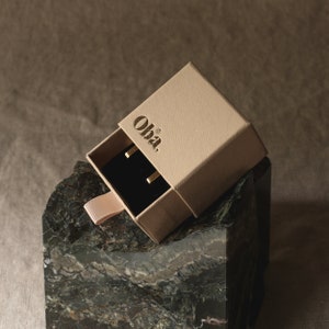 Peut inclure: Une petite bo&icirc;te beige avec le mot "Oha." imprim&eacute; sur le dessus. La bo&icirc;te est ouverte, r&eacute;v&eacute;lant un int&eacute;rieur en velours noir avec deux boucles d'oreilles en or.