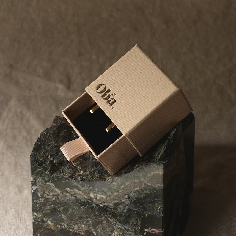 Peut inclure: Une petite bo&icirc;te beige avec le mot "Oha." imprim&eacute; sur le dessus. La bo&icirc;te est ouverte, r&eacute;v&eacute;lant un int&eacute;rieur en velours noir avec deux boucles d'oreilles en or.