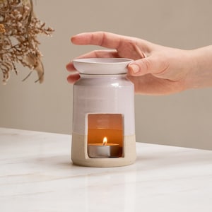 Op de afbeelding: Een witte keramische etherische olieverdamper met een lichtroze glazuur. De diffuser heeft een kleine opening aan de zijkant en een ronde bovenkant. Een klein theelichtje brandt in de diffuser.