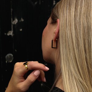 Peut inclure: Une personne portant une boucle d'oreille carr&eacute;e dor&eacute;e et une bague ovale noire.