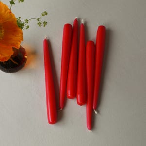 K&ouml;nnte beinhalten: Eine Gruppe von sieben roten Stumpenkerzen, die auf einer wei&szlig;en Oberfl&auml;che lose &uuml;bereinander liegen. Die Kerzen sind alle gleich gro&szlig; und gleich geformt und haben eine spitze Spitze.