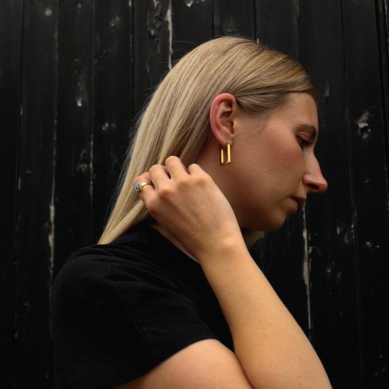 Peut inclure: Une femme aux longs cheveux blonds portant un t-shirt noir et des boucles d'oreilles en or. Elle porte une bague &agrave; son doigt.