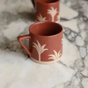 May include: A terracotta mug with a white palm tree design. The mug has a speckled finish and a rounded handle.