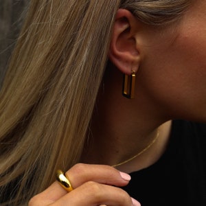 Peut inclure: Une bague en or avec un design arrondi et lisse. La bague est port&eacute;e &agrave; l'index d'une main. La personne porte &eacute;galement des boucles d'oreilles cr&eacute;oles en or.