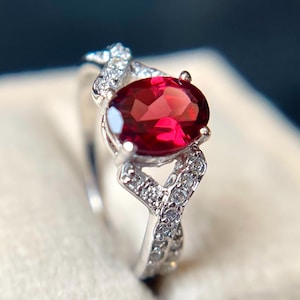 Könnte beinhalten: Ein silberner Ring mit einem großen, ovalen, tiefroten Edelstein. Das Band weist ein verdrehtes Design auf, das mit kleineren, klaren Edelsteinen besetzt ist. Der Ring ist vor einem weichen, cremefarbenen Hintergrund platziert.