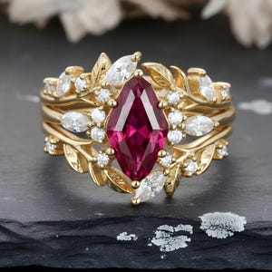 May include: A gold ring set featuring a marquise-cut ruby gemstone as the centerpiece, surrounded by smaller white gemstones and leaf-shaped accents. The ring is set on a dark surface.