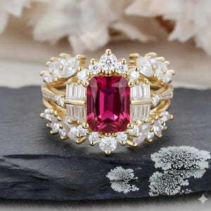 May include: A three-piece gold ring set featuring a large, rectangular, ruby-colored center stone. The ring is surrounded by smaller white and clear stones. The set includes a band with smaller stones and a decorative ring with a floral design.