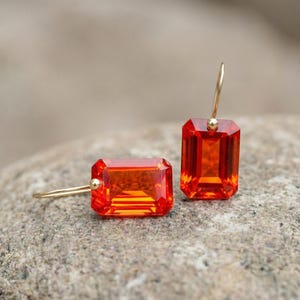 May include: A pair of gold drop earrings featuring rectangular, faceted orange gemstones. The earrings have a simple design with a gold hook and a small gold bead at the top of each gemstone. The gemstones have a vibrant, fiery color.