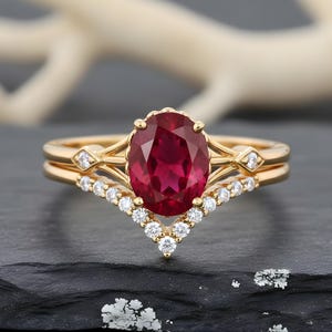 May include: A gold ring set with an oval ruby and a diamond-set band. The ring is paired with a chevron-shaped band, also with diamonds. The design is classic, highlighting the ruby's vibrant red colour. The set is suitable for special occasions.