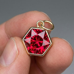 May include: A hexagonal pendant with a vibrant red gemstone. The gemstone is set in a gold-colored frame and has a starburst pattern. The pendant has a small gold loop for a chain.