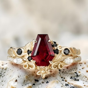 Könnte beinhalten: Ein goldener Ringsatz mit einem großen, facettierten, tiefroten Edelstein als Mittelpunkt. Der Ring ist mit schwarzen und weißen Akzentsteinen sowie floralen und Blattmotiven verziert. Das Band ist ebenfalls goldfarben und mit kleinen weißen Steinen besetzt.