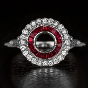 May include: A silver ring with a circular design, featuring a central circle of red gemstones and a halo of white gemstones. The ring has an ornate design and is set against a black background. The ring's design is intricate.