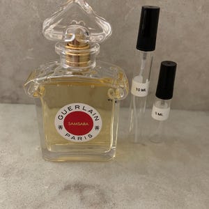 May include: A clear glass Guerlain Samsara perfume bottle with a heart-shaped stopper and gold neck, accompanied by two small clear glass spray vials with black caps, labeled 10 ml and 5 ml, containing a light yellow liquid fragrance.
