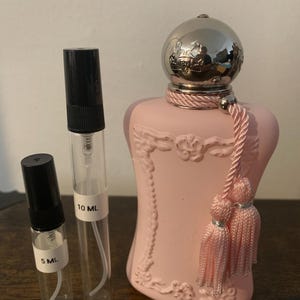 May include: A pink perfume bottle with a silver cap and pink tassels, next to two clear glass vials labeled 5 ML and 10 ML. The perfume bottle has an ornate design.