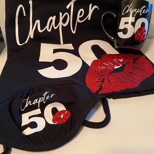 Peut inclure: Ensemble noir avec l'inscription "Chapter 50" et une bouche rouge pailletée. L'ensemble comprend un t-shirt, une tasse à café et un masque facial. Le texte et la bouche sont en blanc et rouge.