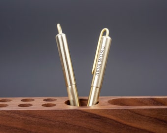 May include: Two gold-toned pens with a brushed finish are standing upright in a wooden pen holder. The pen on the right has the name "ARVA WONG" engraved on the side.