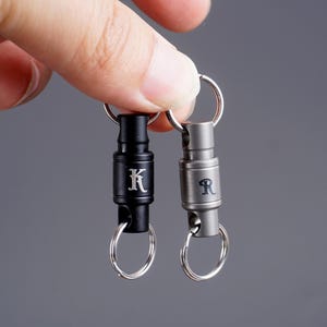 May include: Two metal keychains, one black and one silver, each with a silver keyring. The black keychain has the letter "K" engraved on it, and the silver keychain has the letter "R". Both keychains have a cylindrical design.