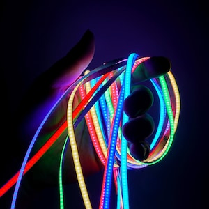 May include: A hand holding a bundle of flexible LED light strips in various colors, including red, blue, green, and yellow. The strips are illuminated, creating a vibrant and colorful display against a dark background.