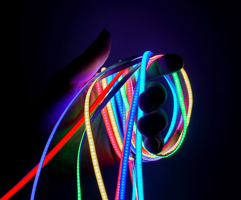 May include: A hand holding a bundle of flexible LED light strips in various colors, including red, blue, green, and yellow. The strips are illuminated, creating a vibrant and colorful display against a dark background.