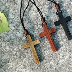 May include: Three wooden cross necklaces with black cords. The crosses are in natural wood tones, dark red, and black. The black cross has the word "VICTORY" engraved on it.