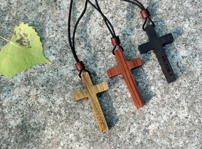May include: Three wooden cross necklaces with black cords. The crosses are in natural wood tones, dark red, and black. The black cross has the word "VICTORY" engraved on it.