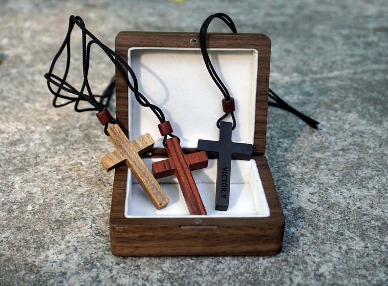 May include: Three wooden cross necklaces in a walnut wood box. The crosses are brown, red, and black. The black cross has the word "VICTORY" engraved on it.