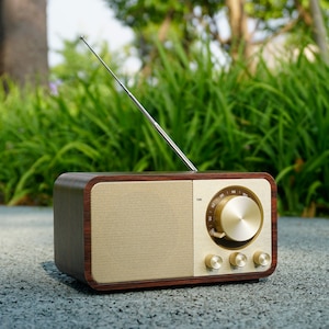 May include: A vintage-style radio with a wooden frame and gold-colored accents. The radio has a speaker, tuning dial, and control knobs. An antenna extends upwards. The radio is set on a gray surface with green foliage in the background.