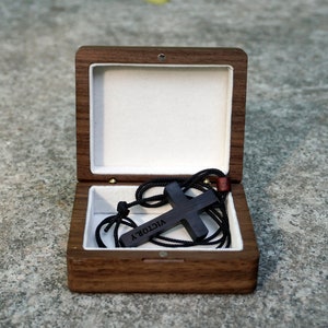 May include: A wooden box with a black wooden cross necklace inside. The necklace has a black cord and the cross has the word "VICTORY" engraved on it.