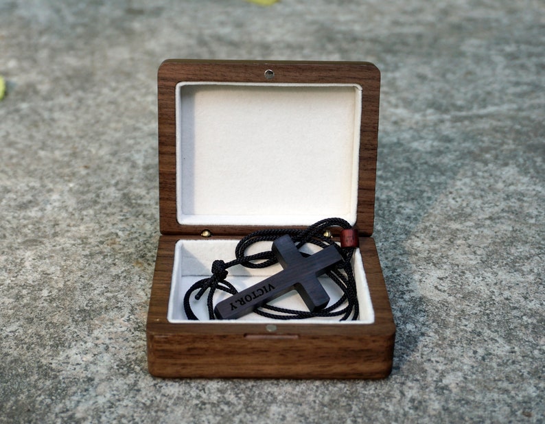 May include: A wooden box with a black wooden cross necklace inside. The necklace has a black cord and the cross has the word "VICTORY" engraved on it.