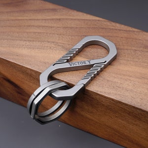 Op de afbeelding: Een zilveren metalen sleutelhanger met een karabijnhaak en een ring. De karabijnhaak heeft het woord "VICTORY" gegraveerd.