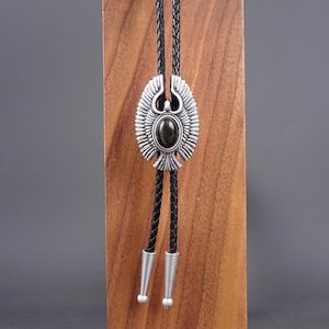 May include: A silver bolo tie with a black leather cord and a silver eagle pendant with a green gemstone in the center.