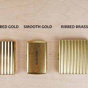 May include: Five metal cigarette cases in silver, gold, brass, and antique finishes. The cases have ribbed or smooth textures, with text labels above each: "Ribbed Silver", "Ribbed Gold", "Smooth Gold", "Ribbed Brass", and "Ribbed Antique".