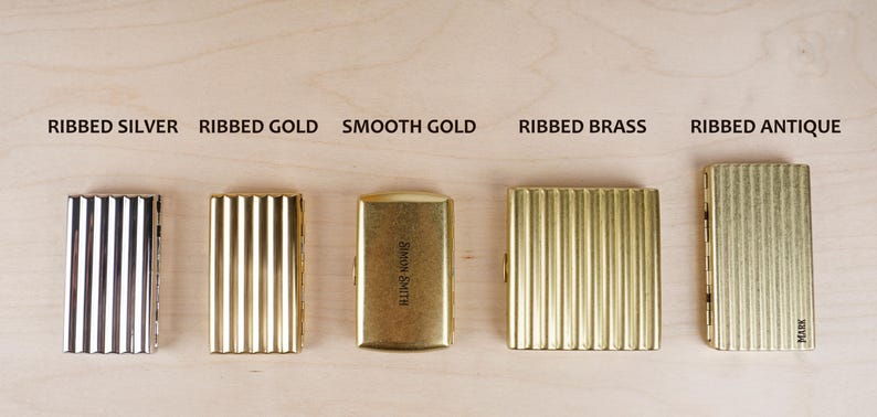 May include: Five metal cigarette cases in silver, gold, brass, and antique finishes. The cases have ribbed or smooth textures, with text labels above each: "Ribbed Silver", "Ribbed Gold", "Smooth Gold", "Ribbed Brass", and "Ribbed Antique".