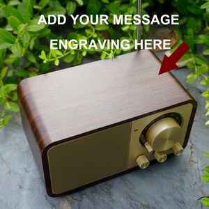 Pu&ograve; includere: Una radio in legno con una griglia per altoparlanti e manopole dorate. La parte superiore della radio &egrave; in legno e ha uno spazio per l'incisione. Il testo "ADD YOUR MESSAGE ENGRAVING HERE" &egrave; scritto sopra la radio.