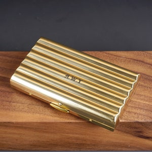 May include: A gold-colored, rectangular case with a ridged design. The case has a clasp closure and the initials "A.W.Y" are engraved on the surface. The case is resting on a wooden surface.