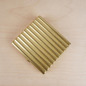 May include: A gold-colored, rectangular compact with a ribbed, textured surface. The compact is positioned diagonally on a light-colored wooden surface. The design features a series of parallel lines across the surface.