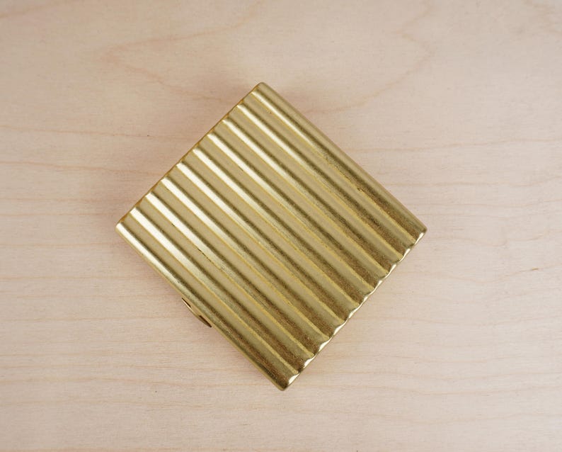 May include: A gold-colored, rectangular compact with a ribbed, textured surface. The compact is positioned diagonally on a light-colored wooden surface. The design features a series of parallel lines across the surface.