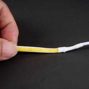 May include: A close-up of a flexible LED strip with a yellow light-emitting surface. The strip is held by a hand, showing the connection to a white cable. The background is a dark surface.