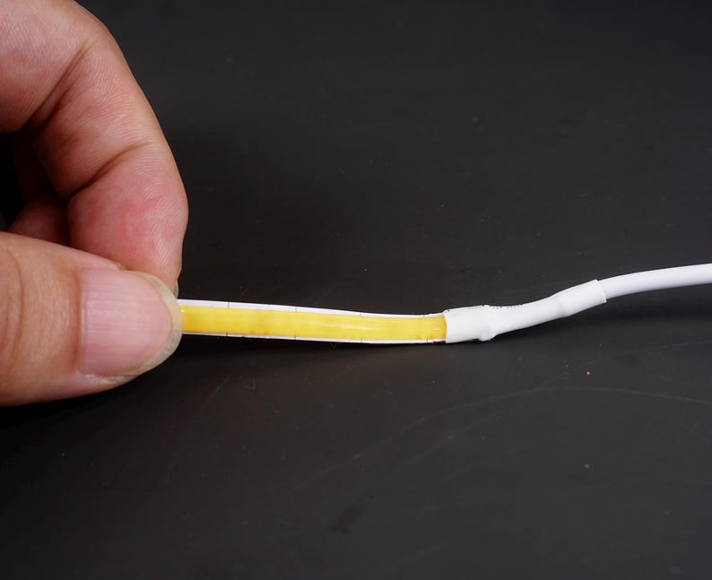 May include: A close-up of a flexible LED strip with a yellow light-emitting surface. The strip is held by a hand, showing the connection to a white cable. The background is a dark surface.