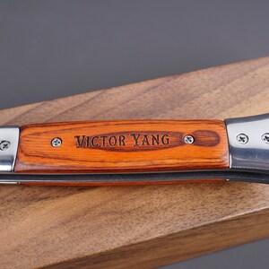 May include: A brown wooden handled pocket knife with a silver blade and the name "VICTOR YANG" engraved on the handle.