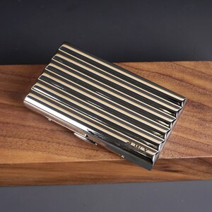 May include: A rectangular, silver-colored metal case with a ribbed design. The case has a shiny, reflective surface and is set against a dark background and a wooden surface. The case appears to be closed.